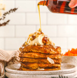 a hand pouring syrup from a jar onto carrot cake pancakes