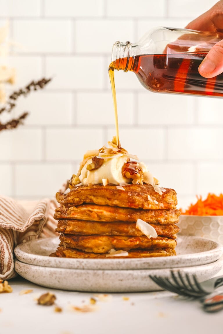 a hand pouring syrup from a jar onto carrot cake pancakes