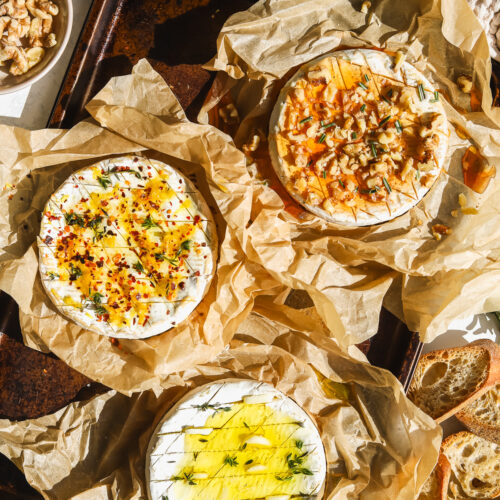 three wheels of camembert cheese, one with lemon and chili flakes, one with garlic and herbs and one with hot honey and walnuts