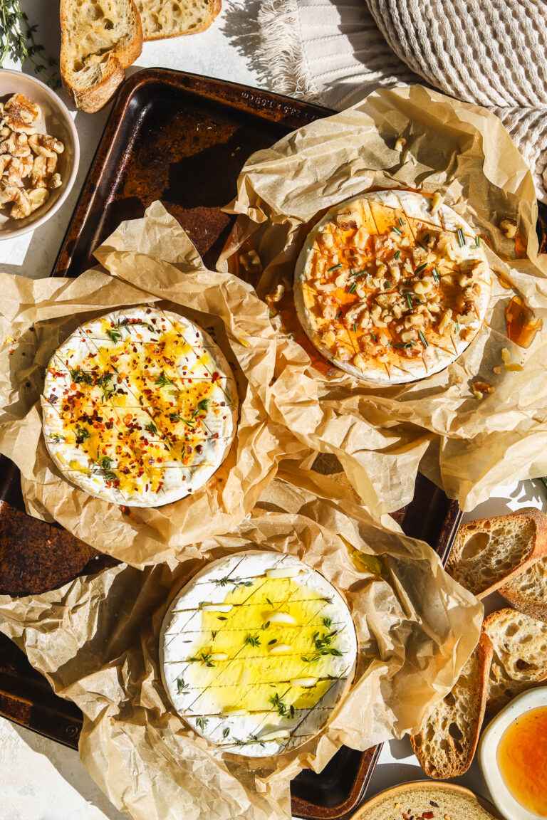 three wheels of camembert cheese, one with lemon and chili flakes, one with garlic and herbs and one with hot honey and walnuts