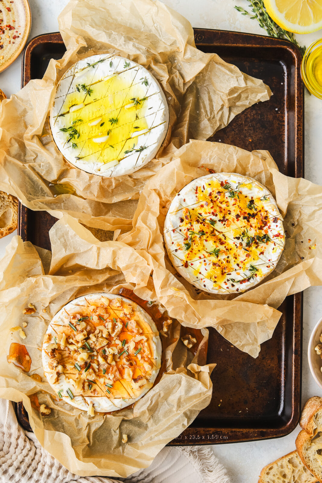 three wheels of camembert cheese, one with lemon and chili flakes, one with garlic and herbs and one with hot honey and walnuts