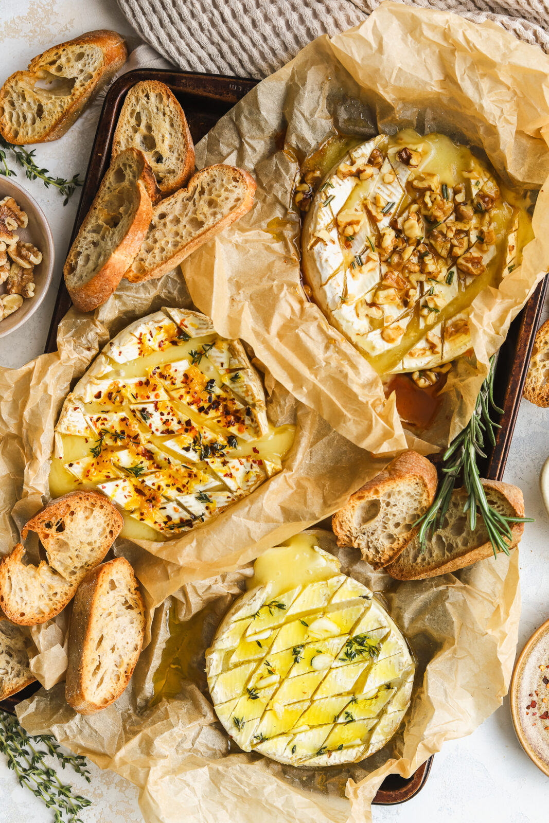 three wheels of baked camembert cheese, one with lemon and chili flakes, one with garlic and herbs and one with hot honey and walnuts