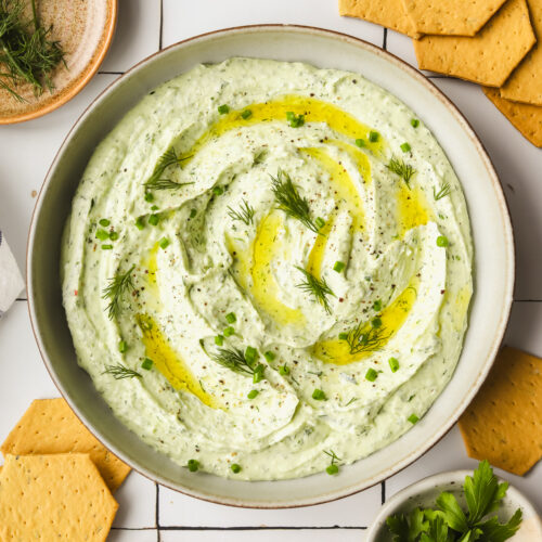 herby goat cheese spread in a shallow bowl with crackers and fresh herbs