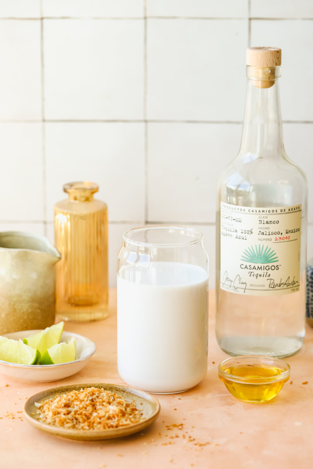 coconut milk in a glass, toasted coconut on a plate, tequila bottle and lime wedges