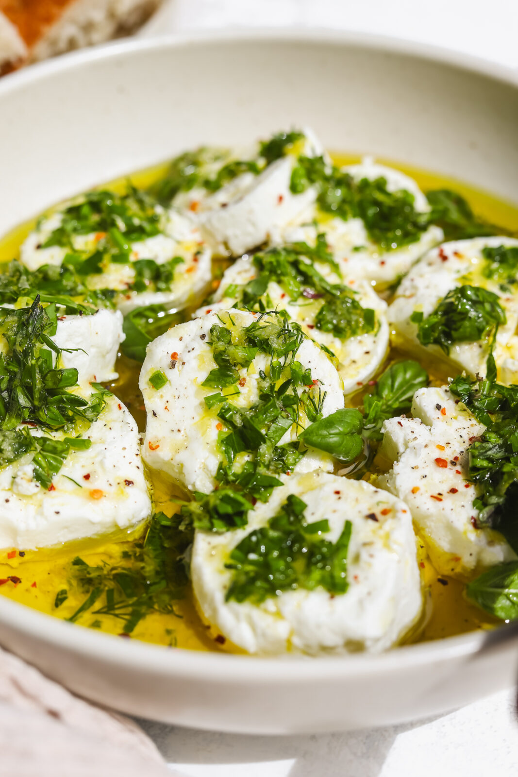 close up of goat cheese rounds in a bowl with fresh herbs and olive oil