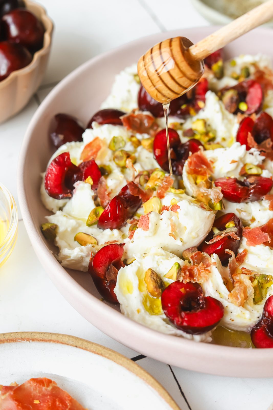 honey being drizzled on a plate of burrata and prosciutto with fresh cherries
