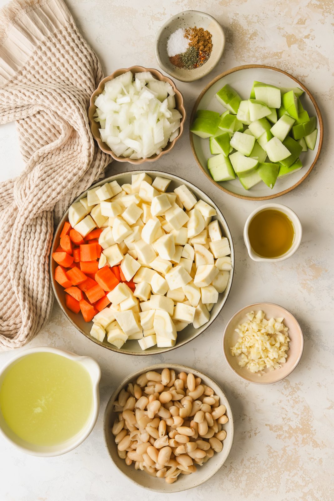 chopped parsnips in a bowl with chopped carrots, plated of chopped apples, plate of white beans, measuring cup of chicken broth, bowl of chopped onions and spices in bowls